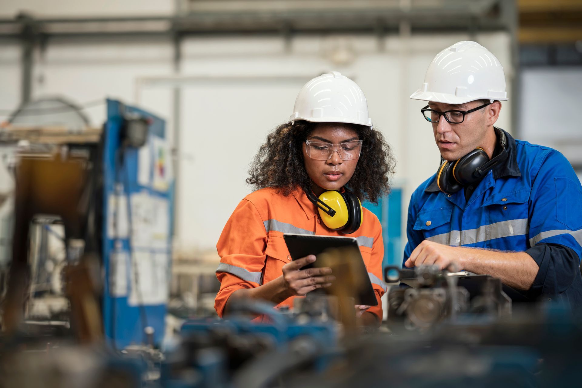Two engineers in hard hats and workwear, inspecting machinery with a tablet.