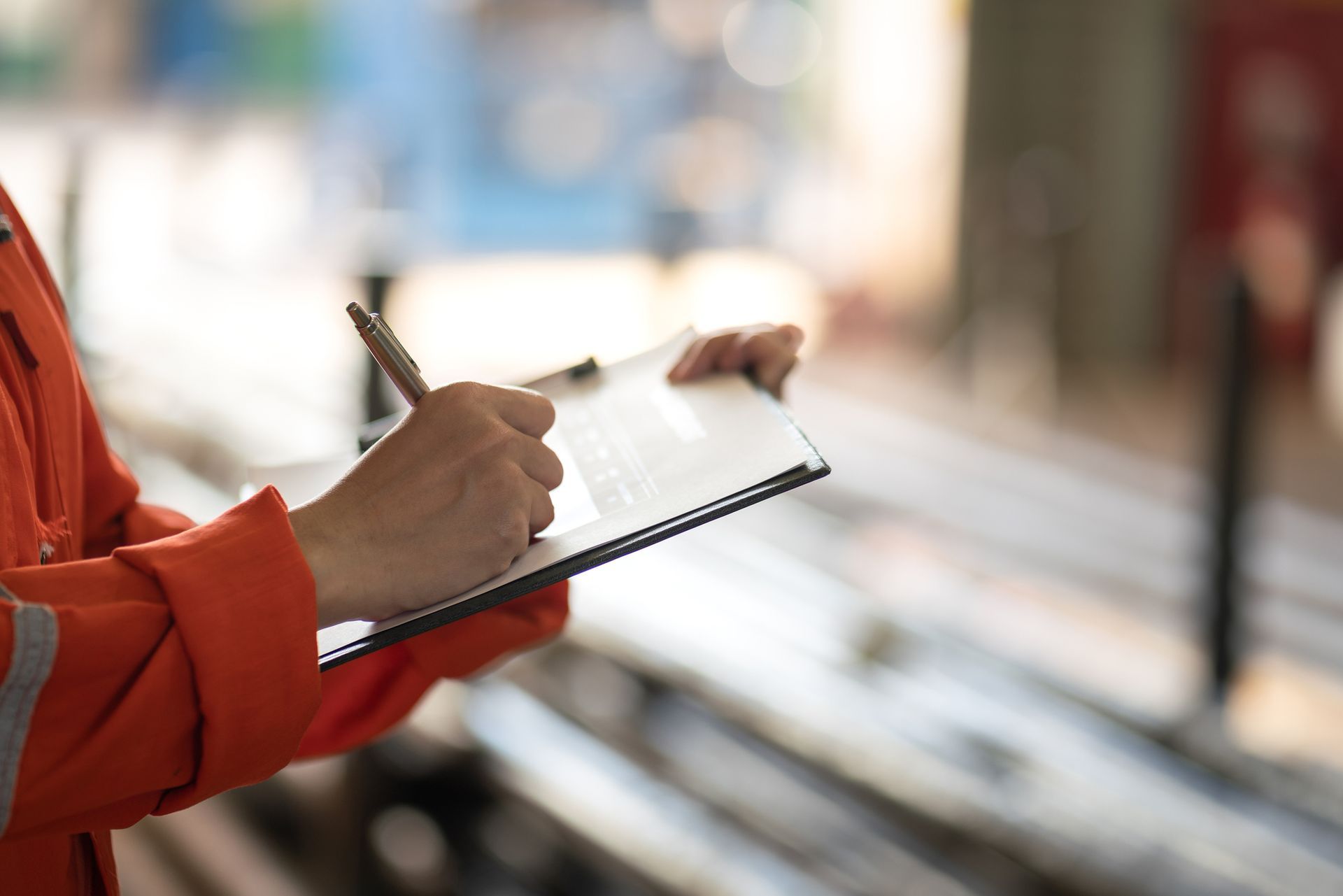 Person in orange work clothes writing on a clipboard, industrial setting.