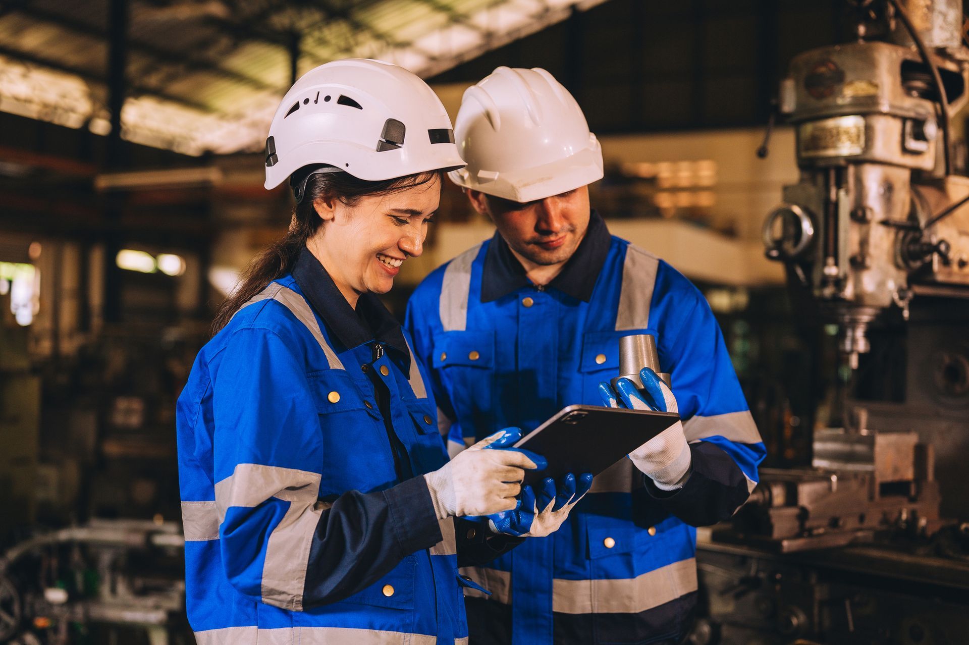 Two workers in blue uniforms and hard hats, looking at a tablet in a factory.