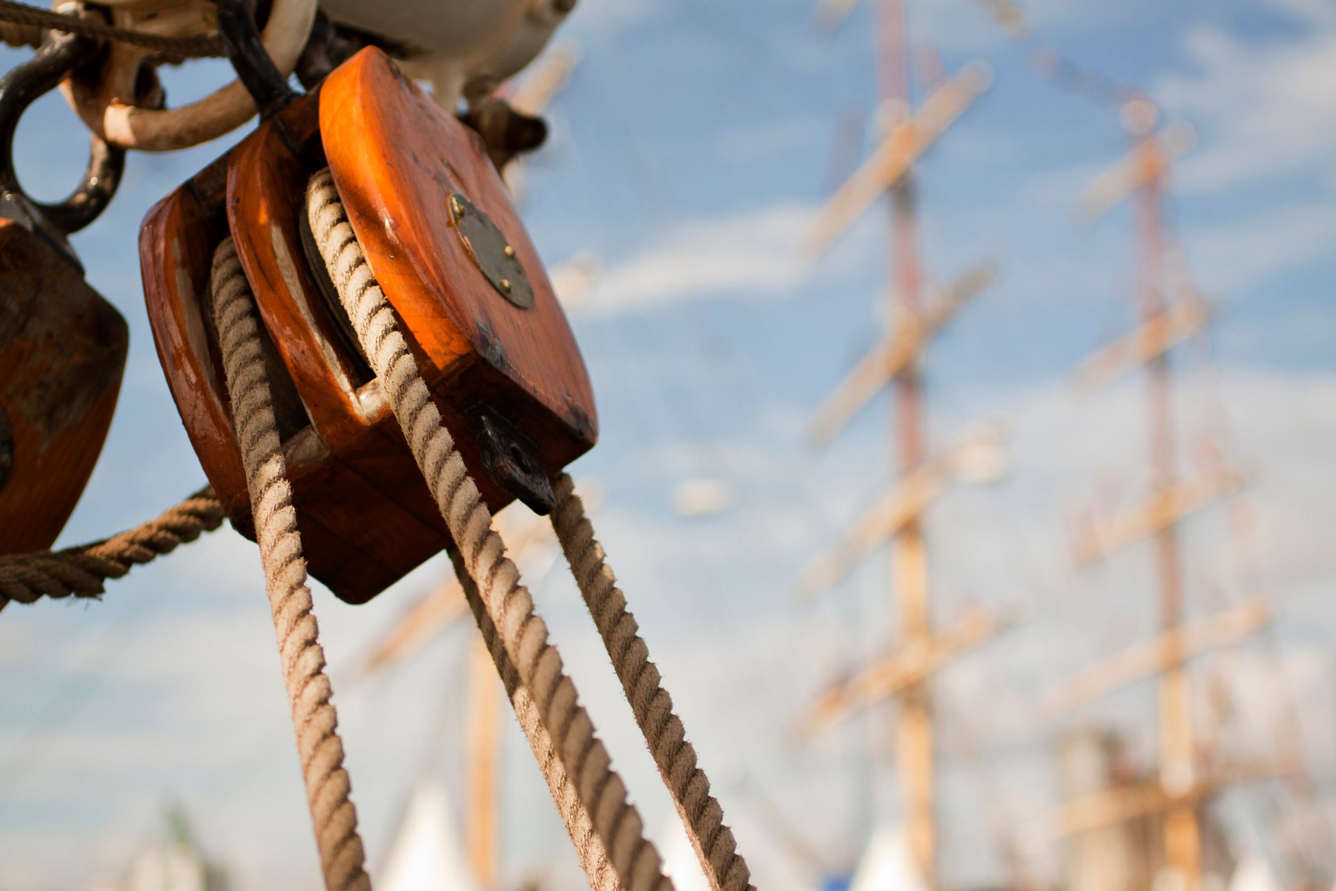 Wooden pulley system with ropes on a sailboat; masts and sky in the background.