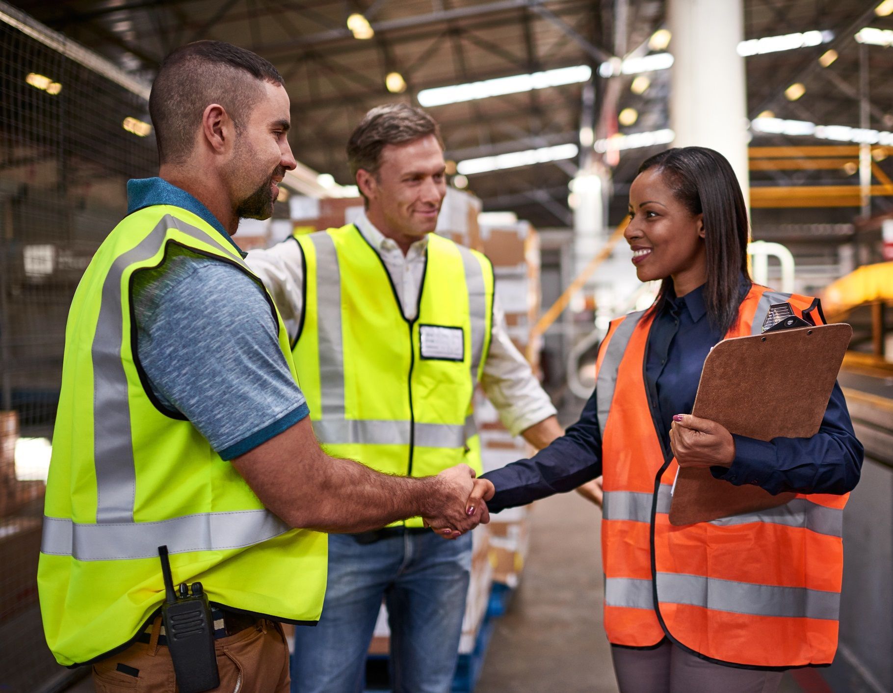 Three people in vests shake hands in a warehouse.