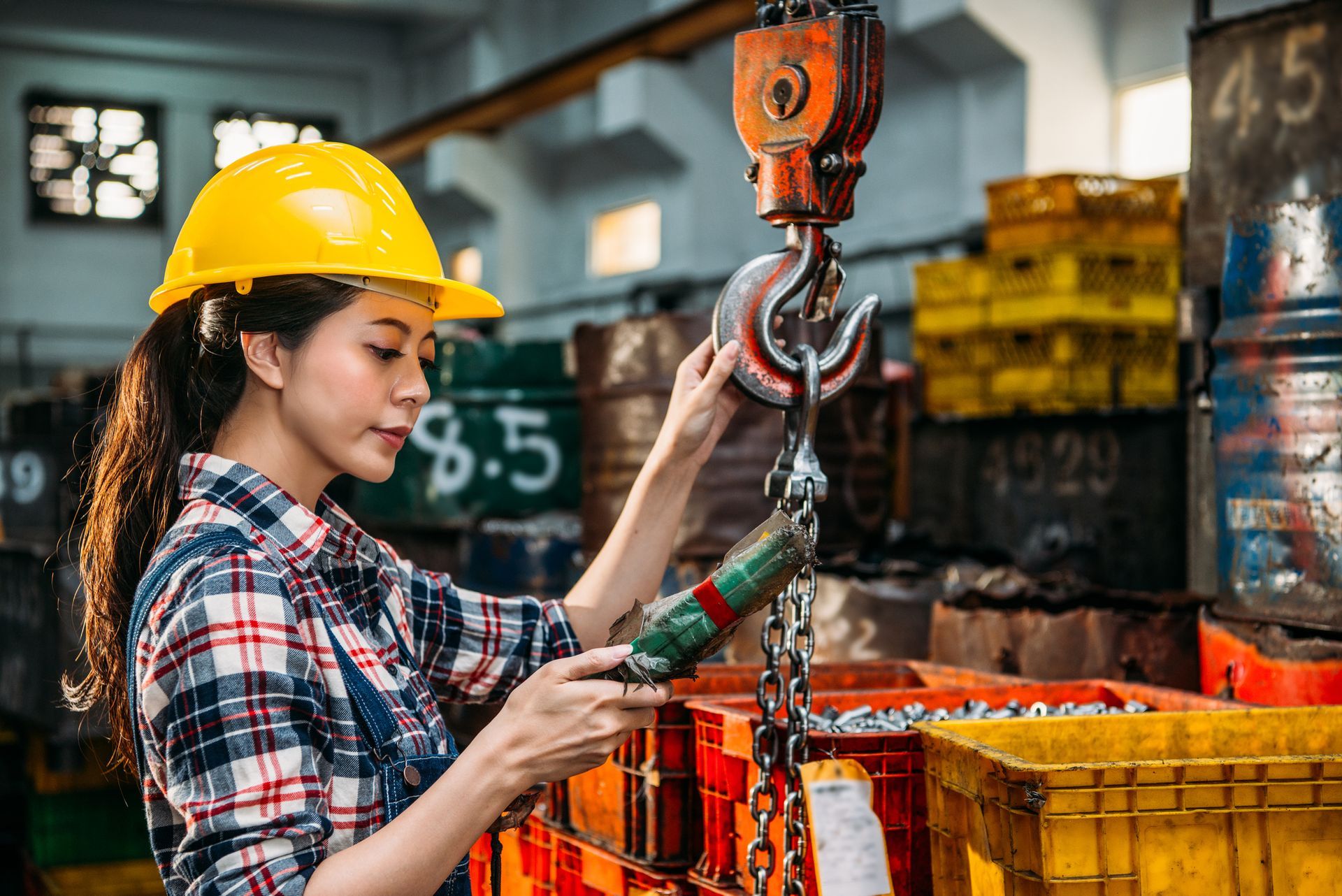 Woman in yellow hard hat inspecting a crane hook in a factory setting.
