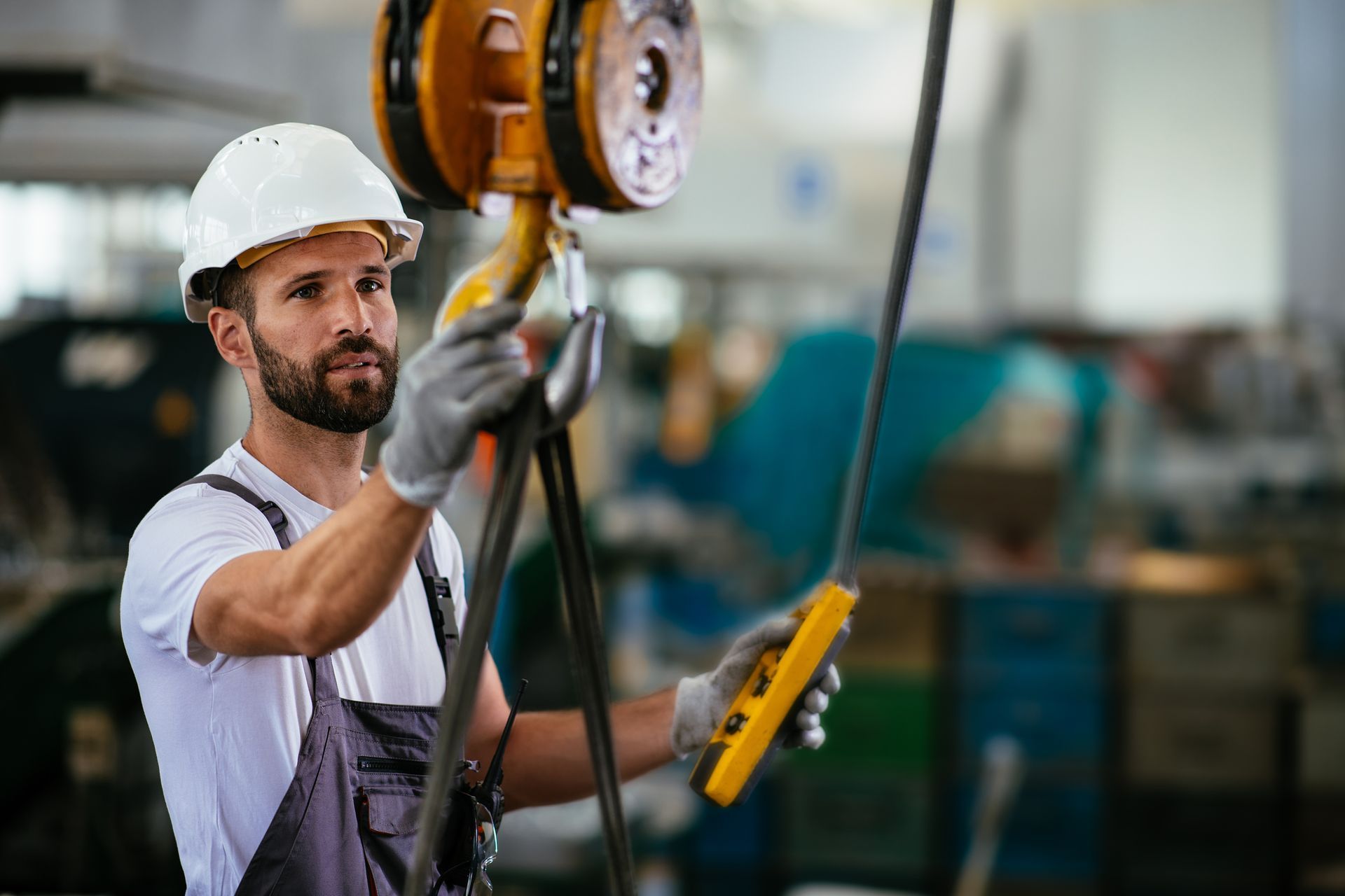 Man in a white hard hat and gloves operating an overhead crane in a factory.