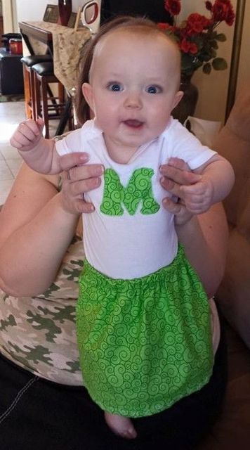 A baby wearing a white bodysuit with a green letter "M" and a green patterned skirt, held up by an adult.