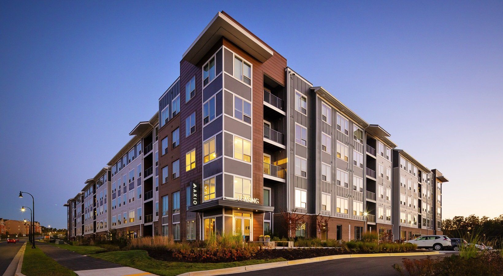 Modern multi-story apartment building with dark gray and brick facade, lit windows, clear blue sky.