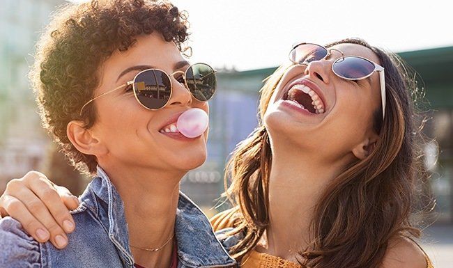 Two women laughing, one with bubble gum, both wearing sunglasses, outdoors.