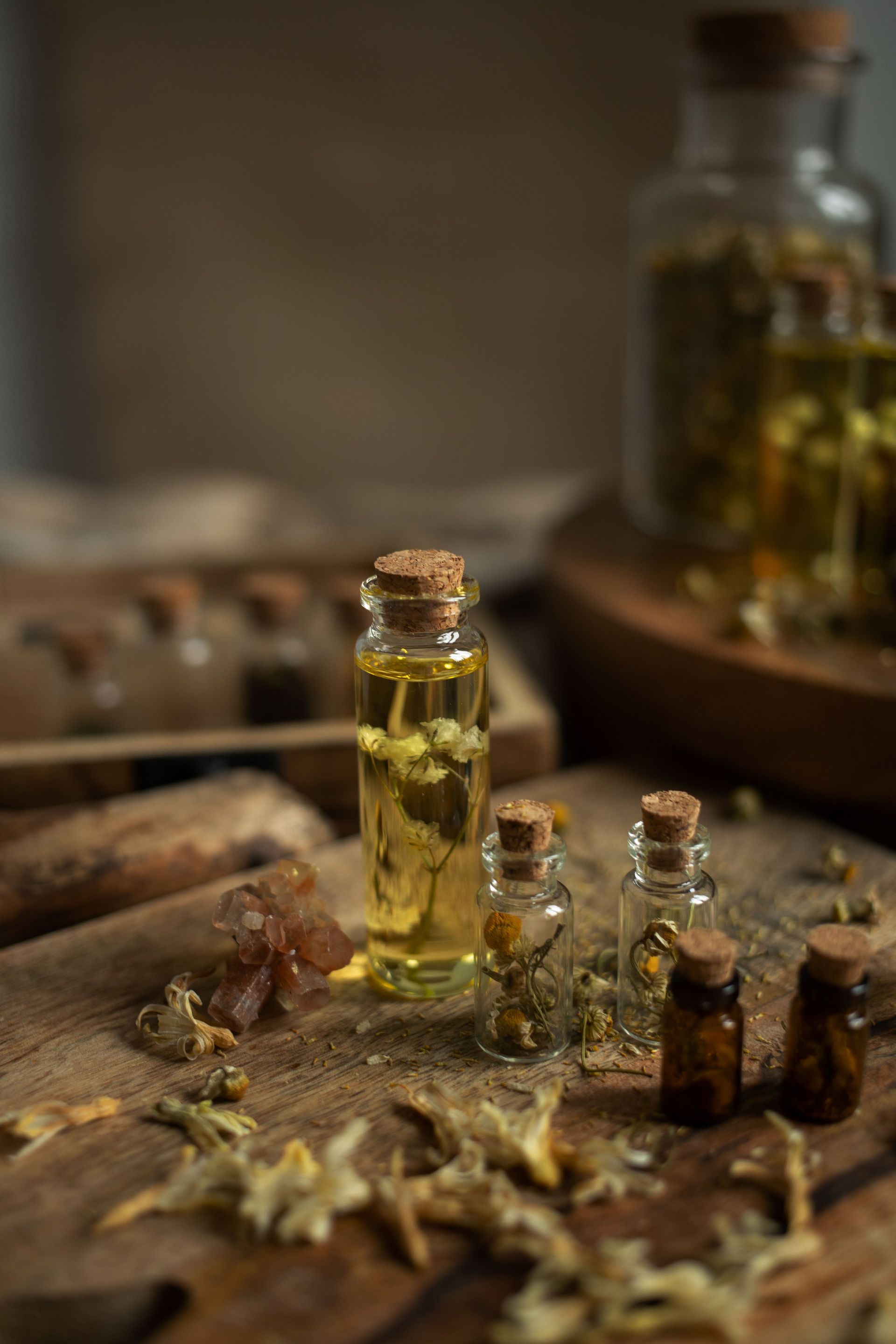 Small glass vials of herbal oil on a wooden surface with dried leaves and a blurred bottle in the background