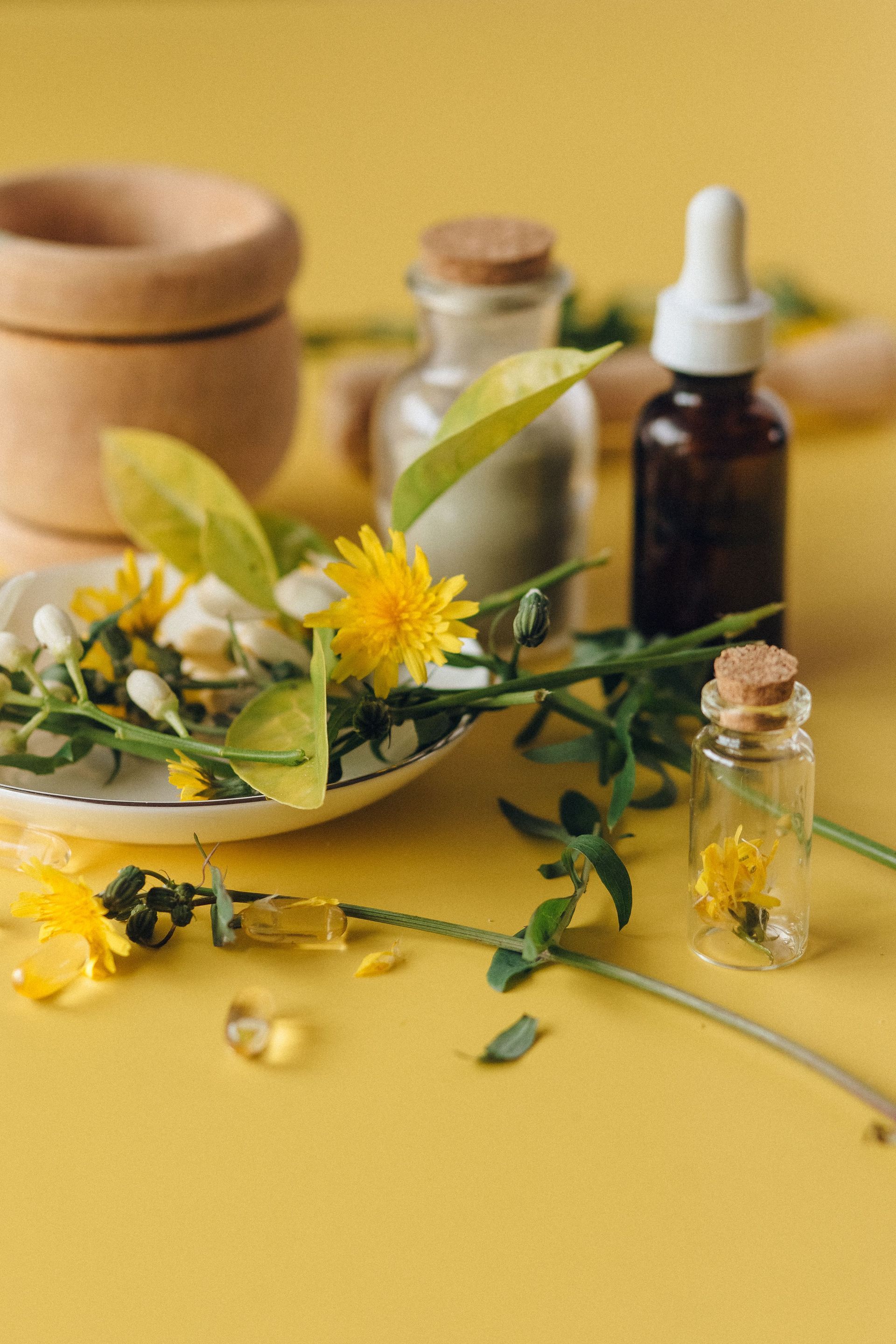 Natural skincare still life with yellow flowers, small glass bottles, and a wooden jar on a yellow background