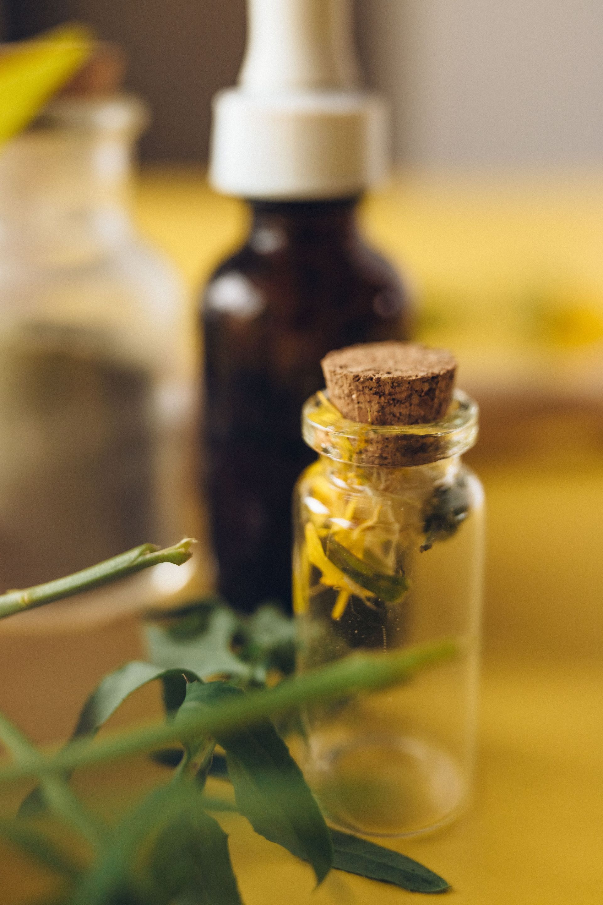 Small glass vial with cork and yellow flowers, with a blurred dark bottle in the background.