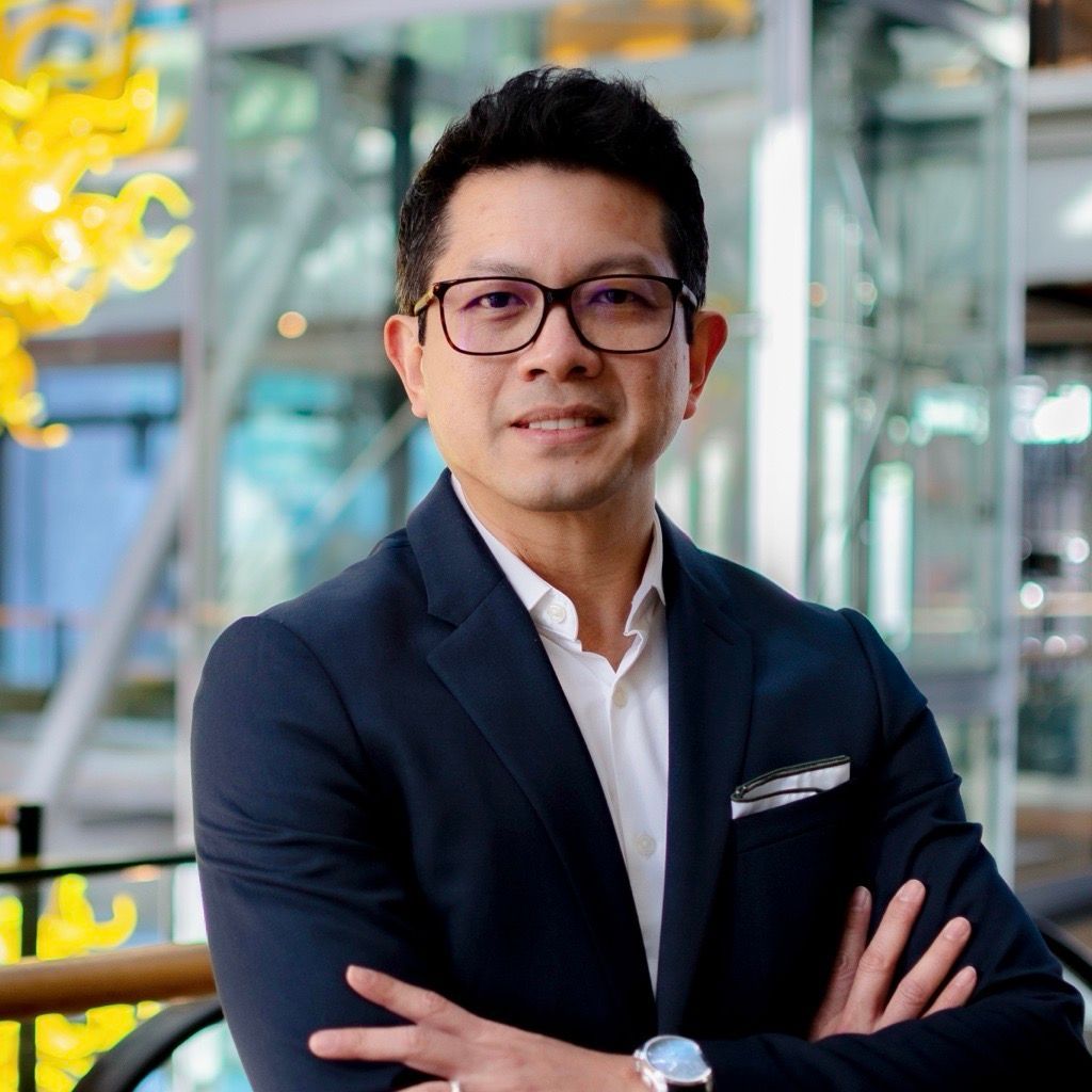 Man in a suit smiles, arms crossed, wearing glasses. Set in a modern building with a glass background.