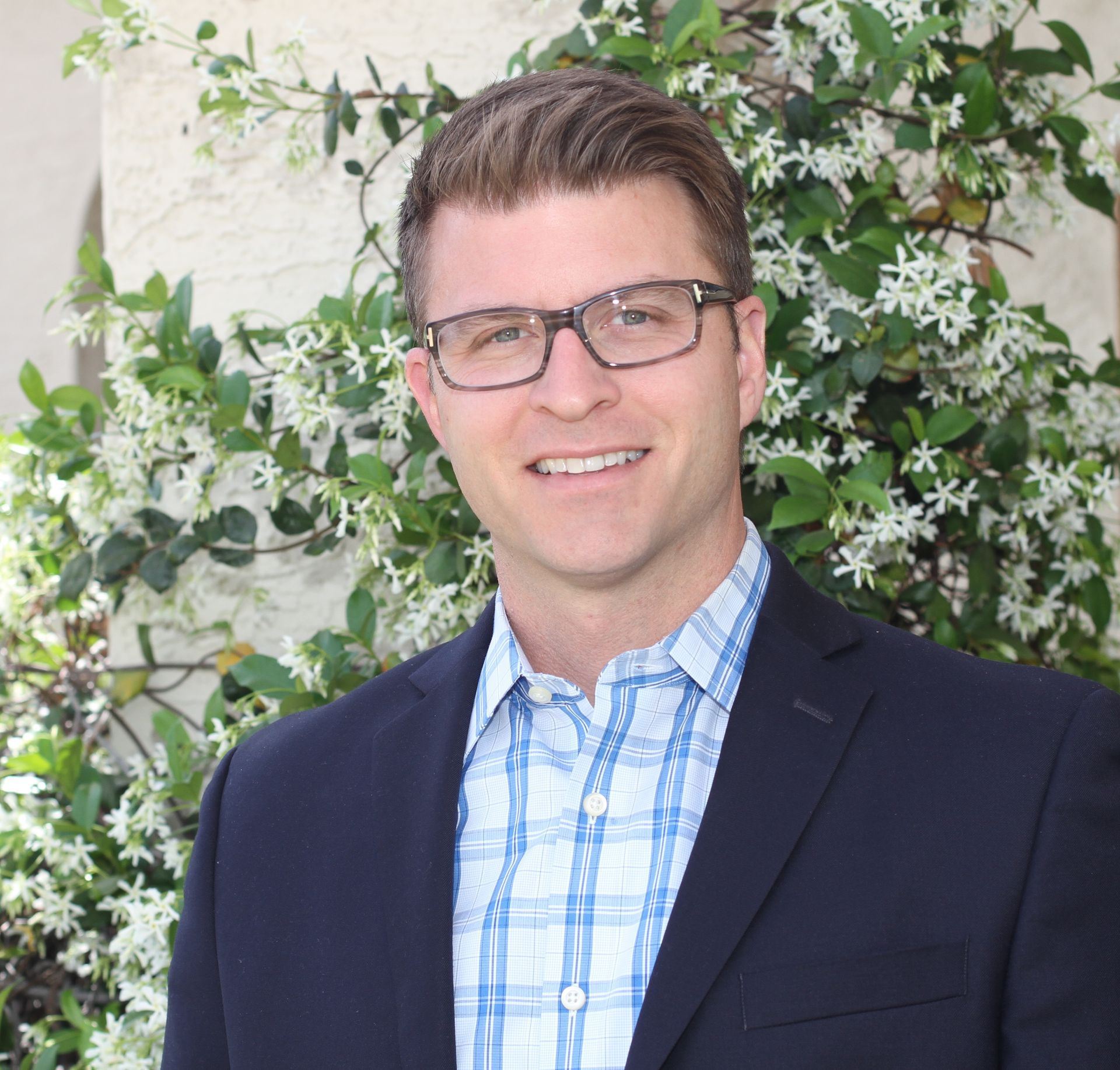 Man in blazer and glasses smiling, standing in front of white flowers and green leaves.