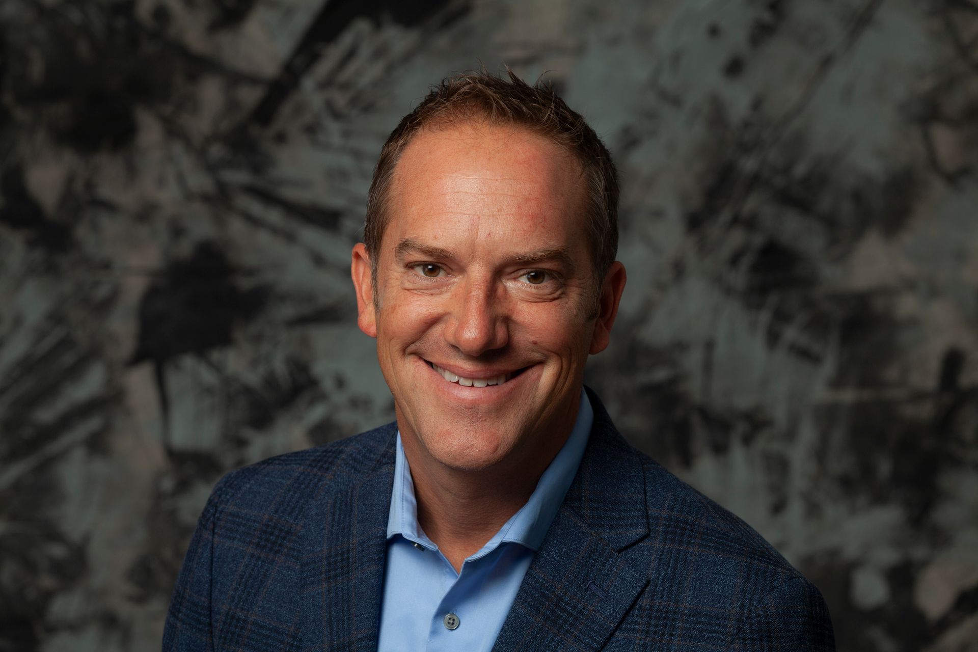 Man in a blue blazer and button-down shirt smiles in front of a textured background.