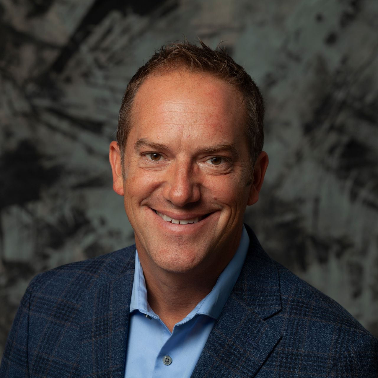 Man in a blue blazer and button-down shirt smiles in front of a textured background.