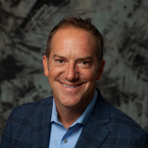 Man in a blue blazer and button-down shirt smiles in front of a textured background.