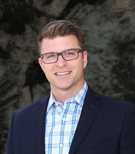 Man in blazer and glasses smiling, standing in front of white flowers and green leaves.