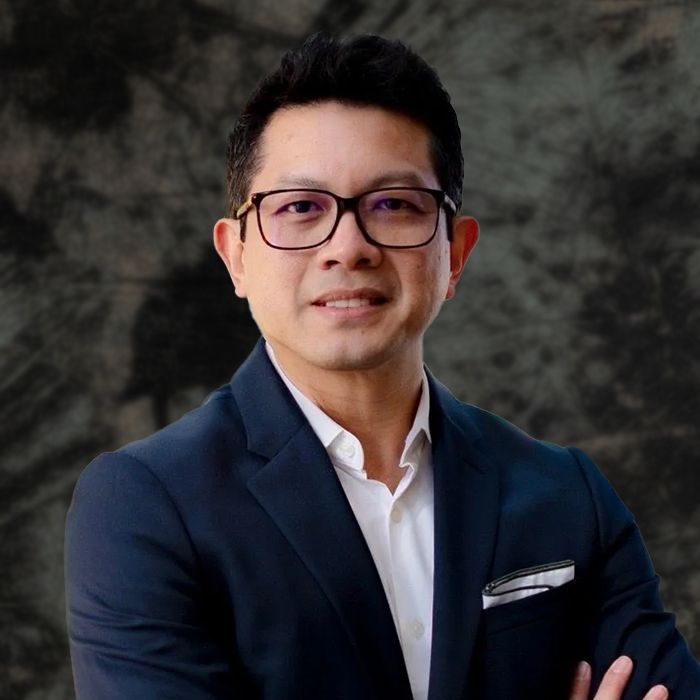 Man in a suit smiles, arms crossed, wearing glasses. Set in a modern building with a glass background.