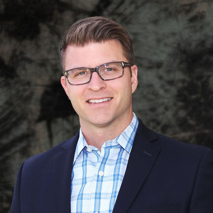 Man in blazer and glasses smiling, standing in front of white flowers and green leaves.