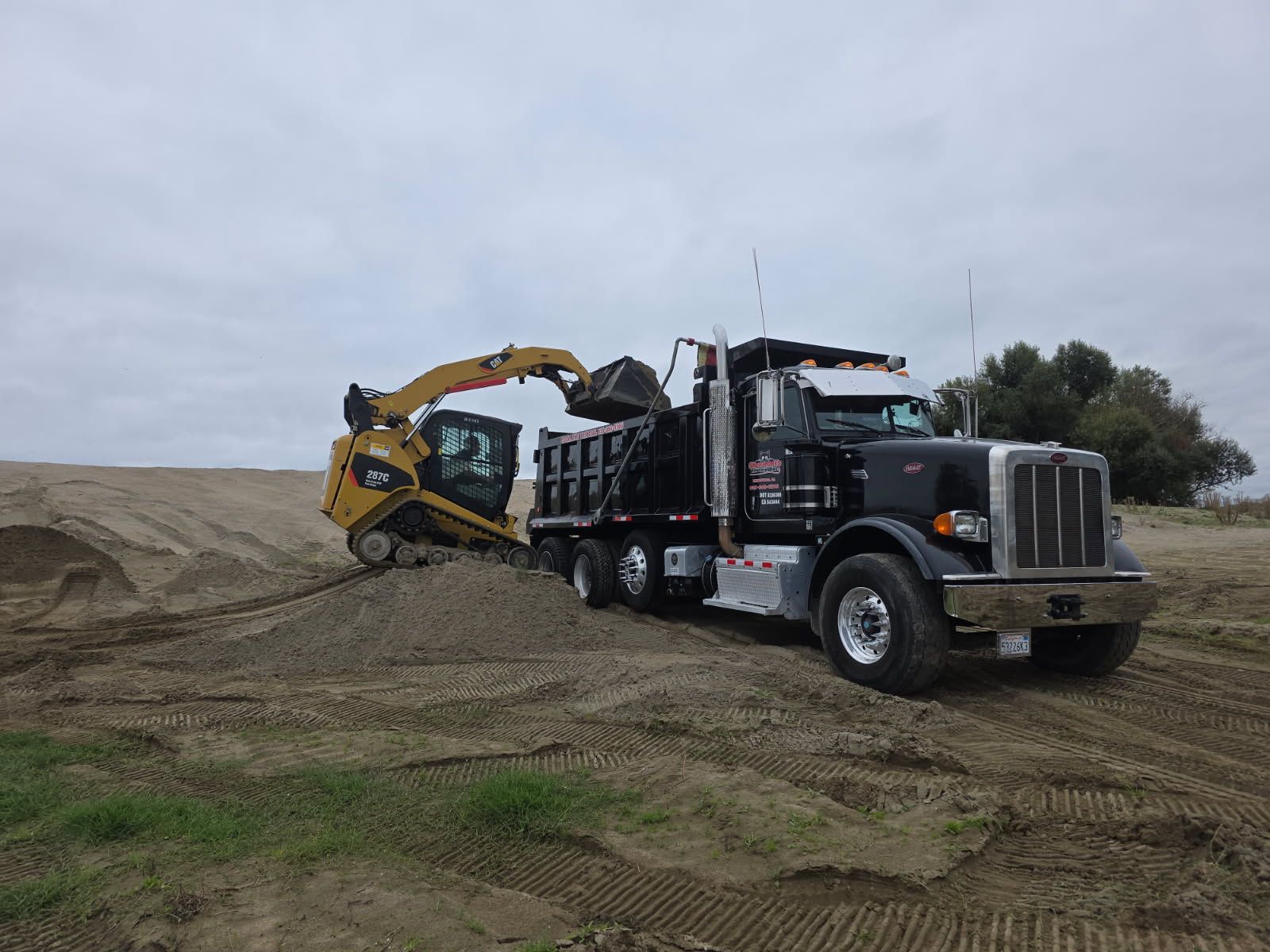 An excavator loads debris into a dump truck, creating dust on a construction site