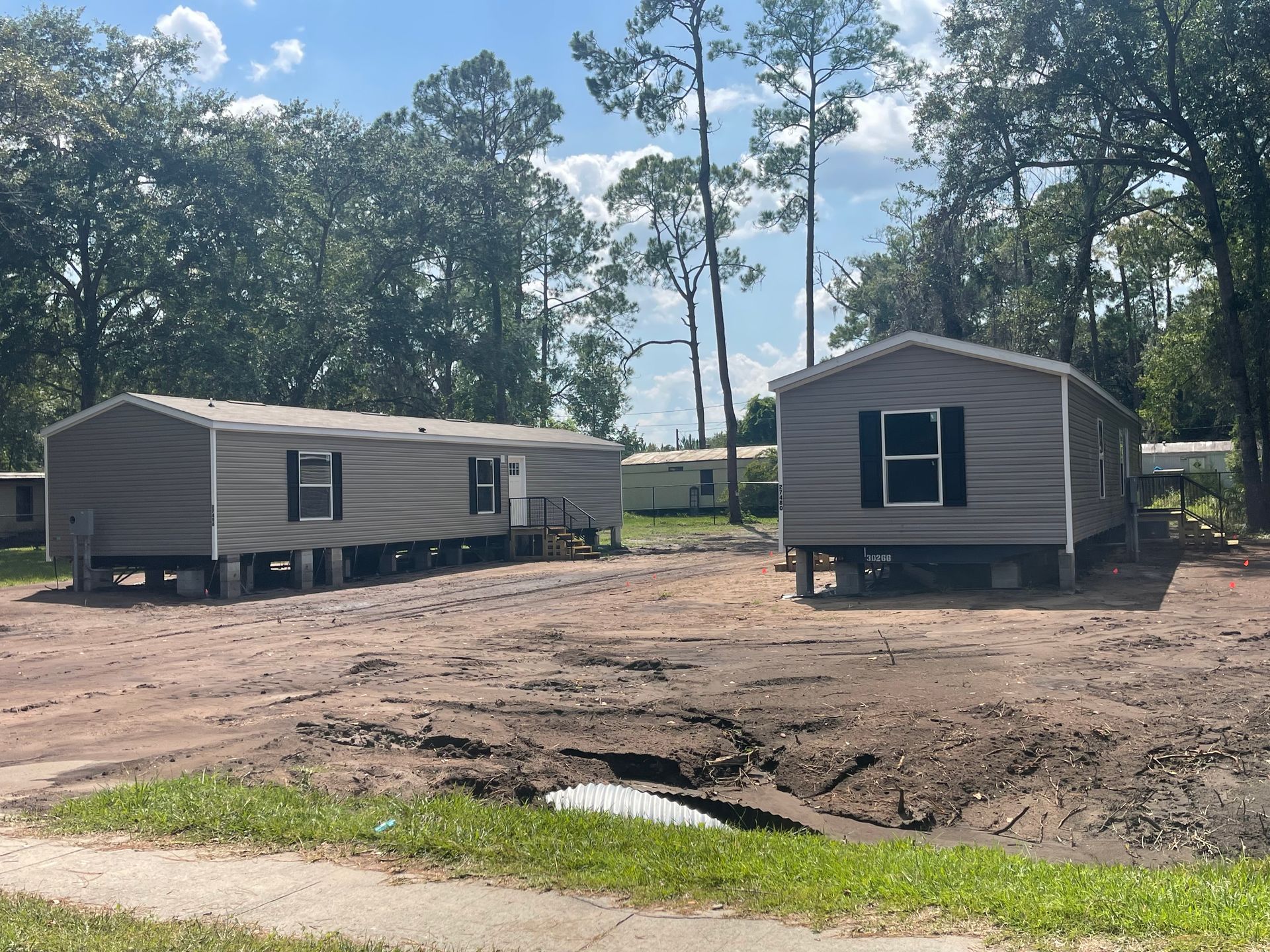 Two mobile homes are parked in a dirt lot with trees in the background