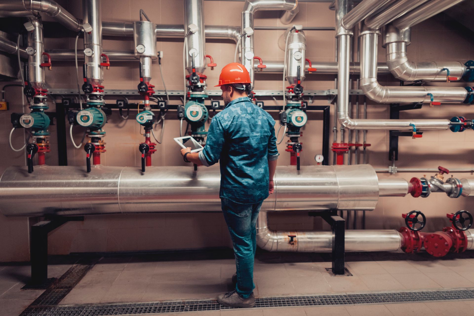 Technician in hard hat inspects pipes in a mechanical room, using a tablet.