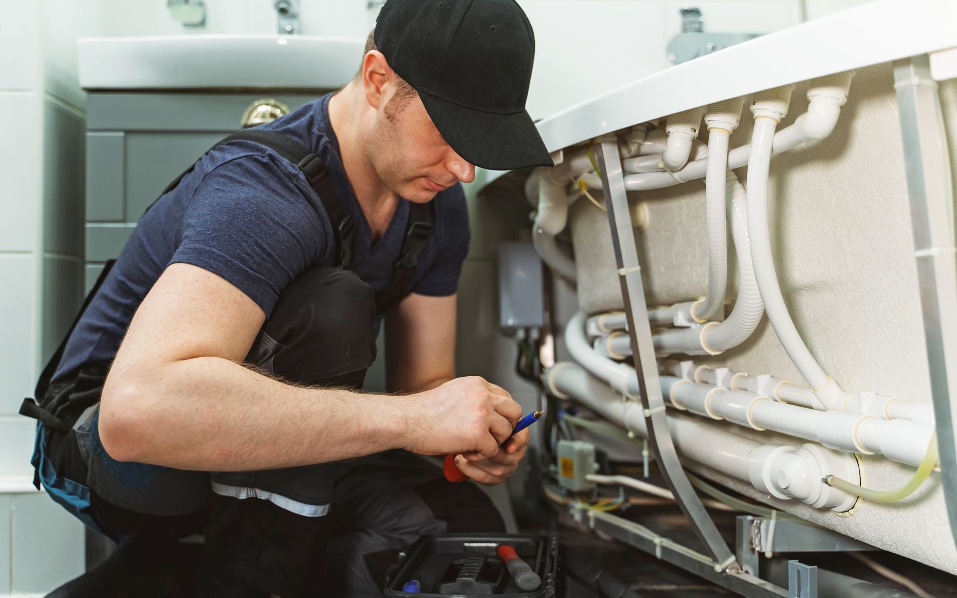 Plumber in a black cap, crouched, working on pipes under a white bathtub.