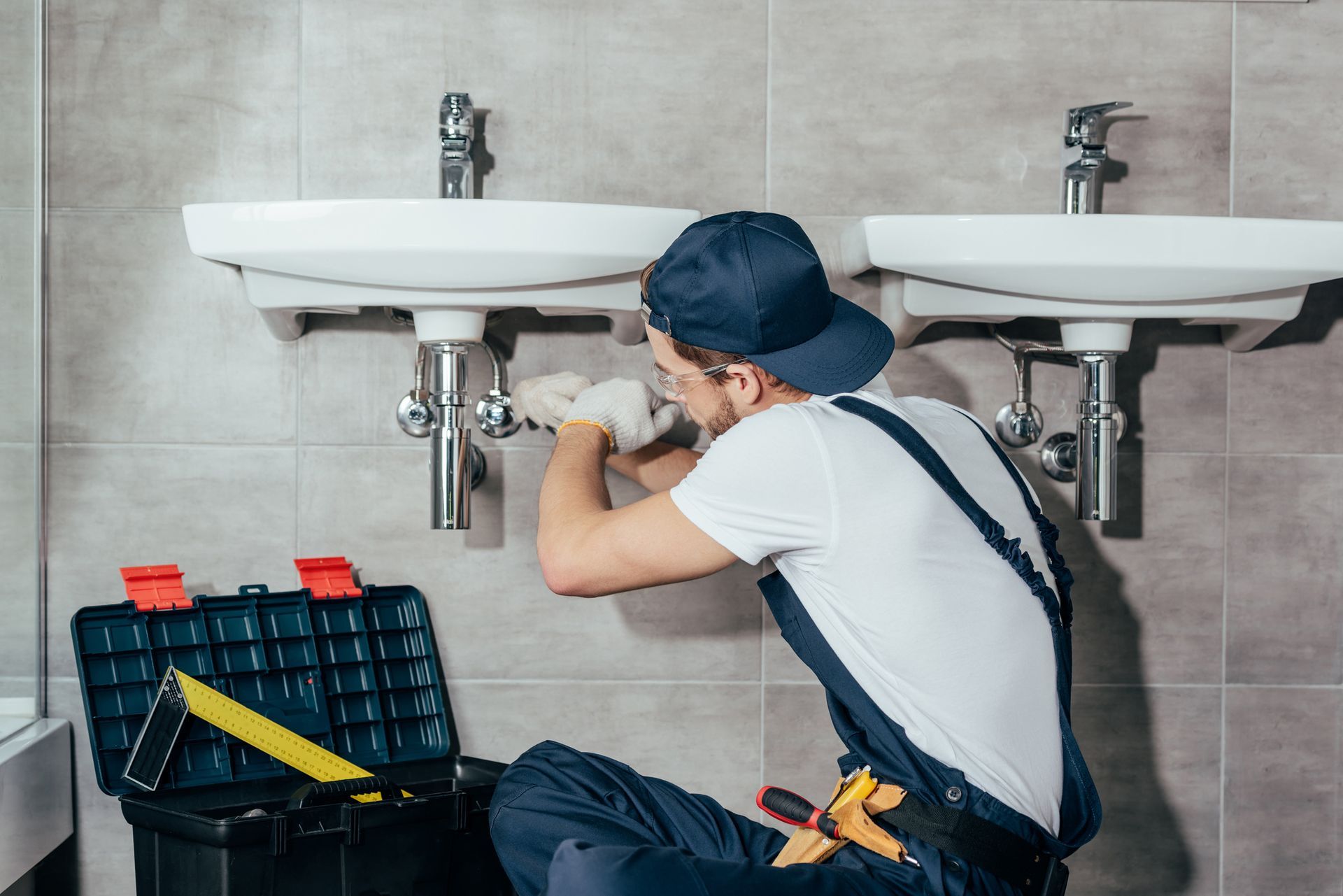 Plumber in a blue cap and overalls repairs a sink in a bathroom with a toolbox nearby.