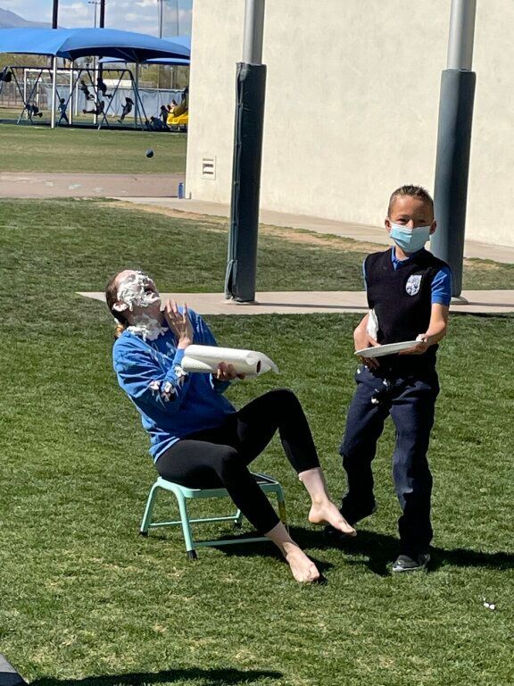 Two people on a grassy field, one seated and laughing while the other stands beside them wearing a mask.