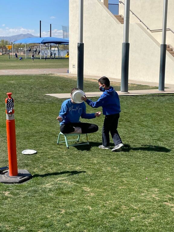 Two people on a grassy field, one kneeling and one standing, reaching toward each other near playground equipment.