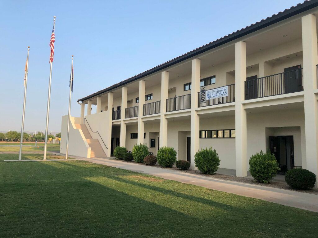 Two-story beige building with columns, flagpoles, and a grassy lawn under a clear sky