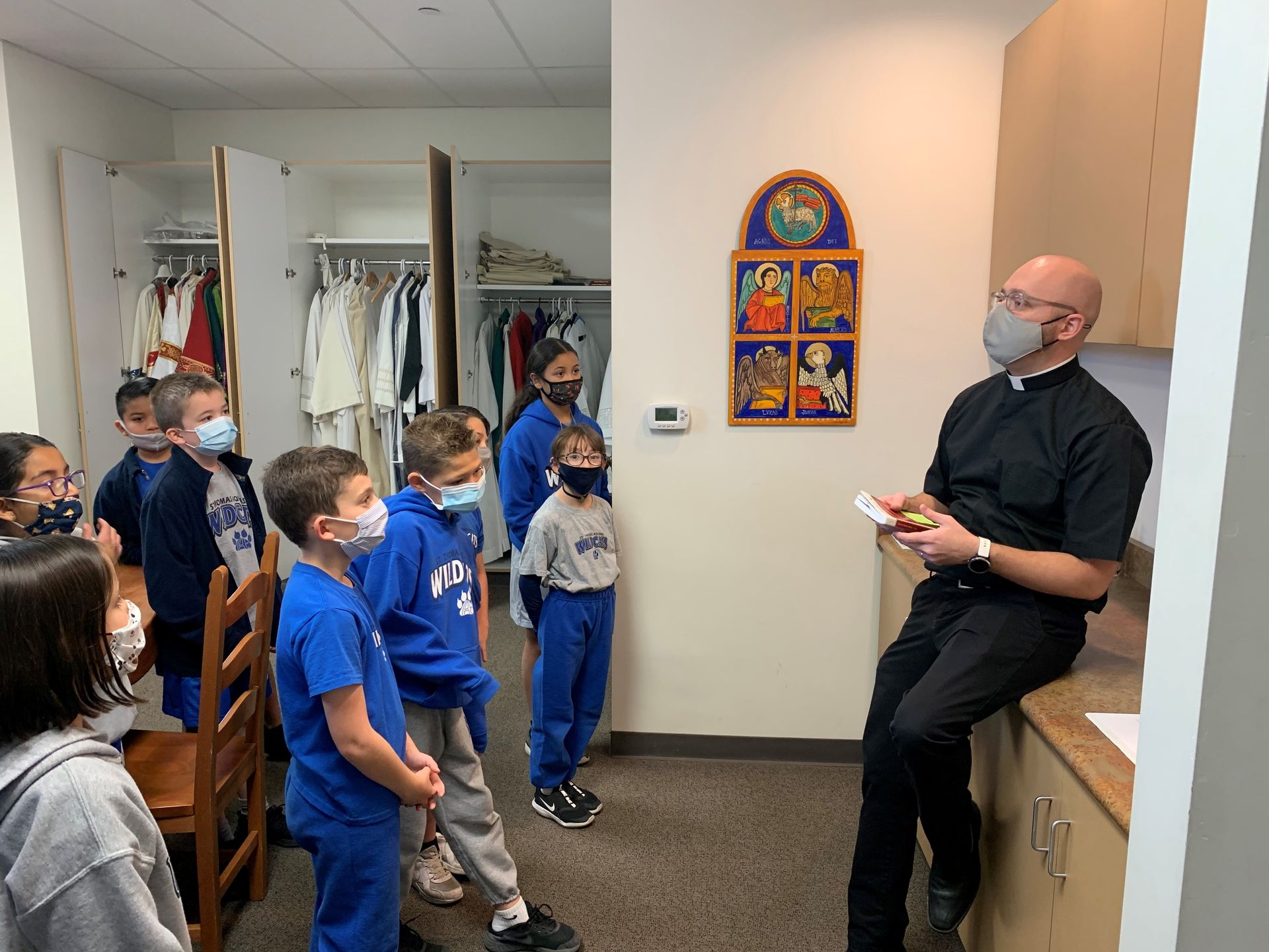 Children in blue uniforms stand in a room while a masked adult sits by cabinets, speaking to them.