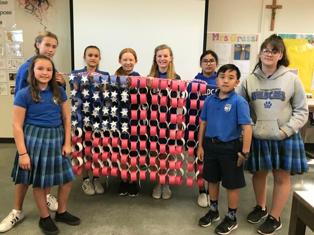 Group of students holding a red, white and blue star-patterned quilt in a classroom.