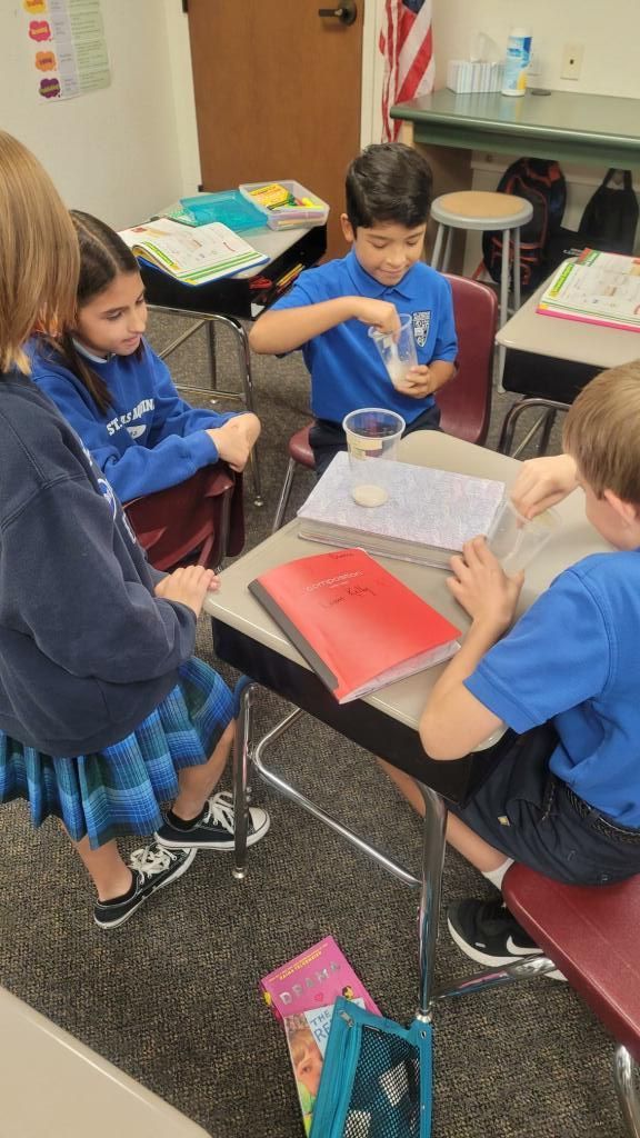 Students in blue uniforms work together at a classroom table with books and art supplies.