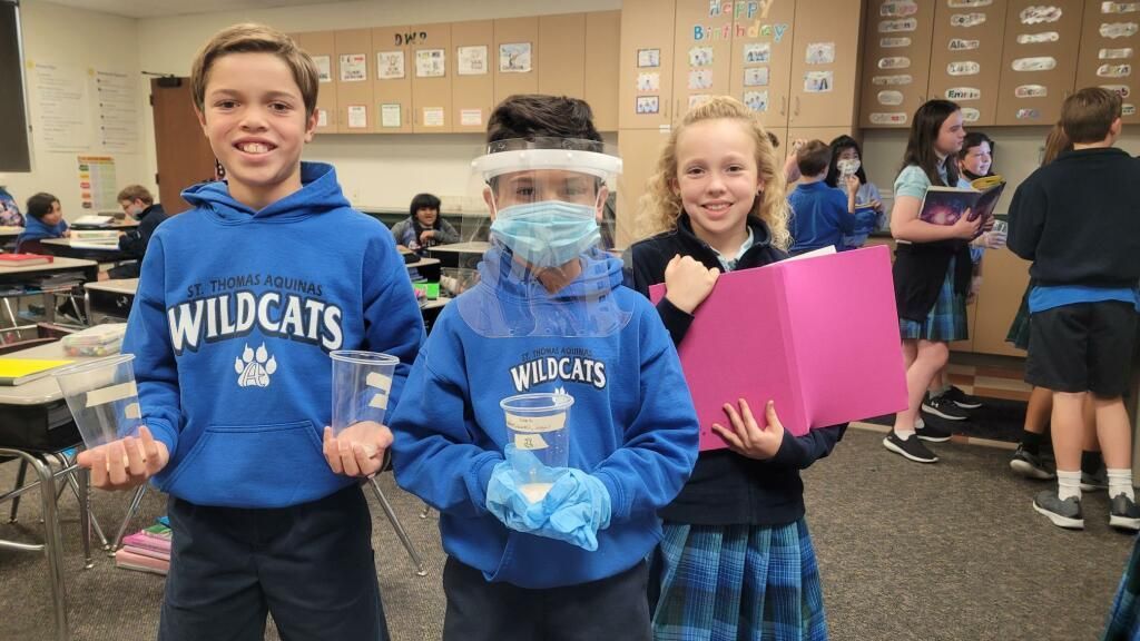 Three students in blue Wildcats hoodies holding items in a classroom, smiling at the camera.