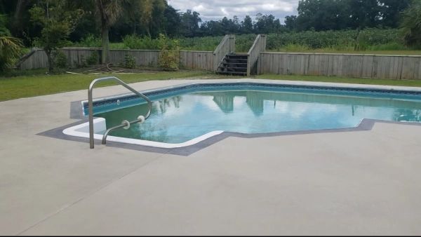 Swimming pool with gray concrete deck, blue water, and wooden steps leading up to a wooden fence.