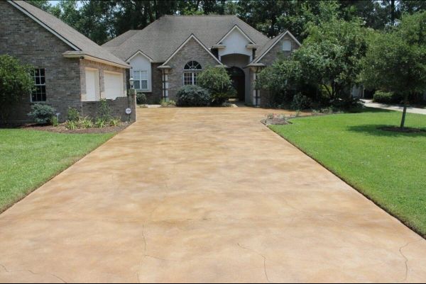 Tan concrete driveway leading to a brick home with manicured lawn and landscaping.