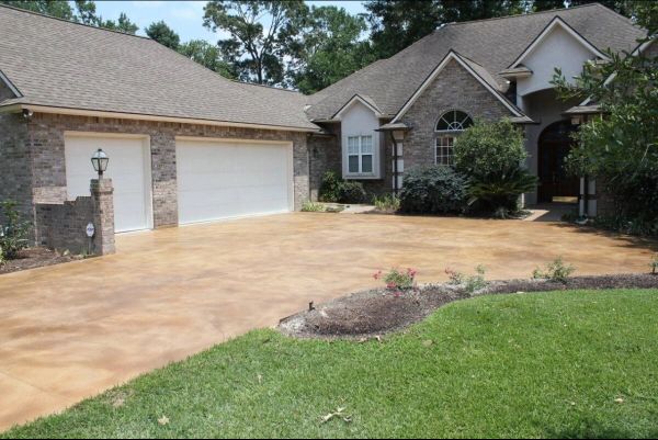 Tan-colored concrete driveway leading to a brick-faced house with a two-car garage and a green lawn.
