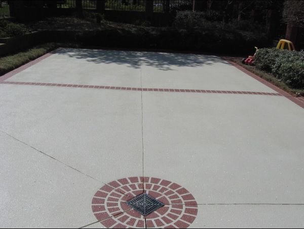 Large, light-colored concrete patio with brick borders and a decorative brick circle. Sunlight.