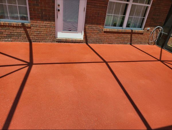 Red concrete patio with shadows cast from a structure, adjacent to a brick wall, door, and windows.
