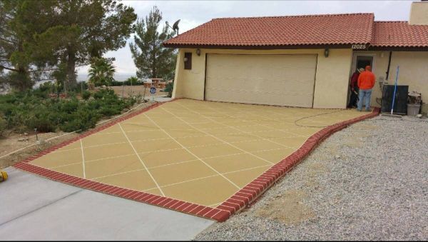 Tan driveway with brick border in front of a house with a closed garage door and two people standing nearby.