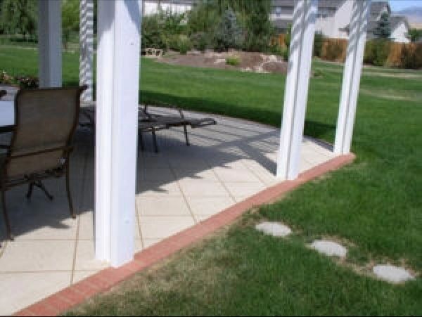 White pergola with tiled patio, red brick border, green lawn, and patio furniture.