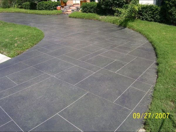 Dark gray concrete driveway with a flagstone pattern, curving through green grass towards a house.