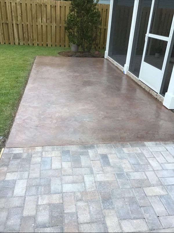 Brown concrete patio next to a brick patio, with a green lawn, wooden fence, and screened porch.