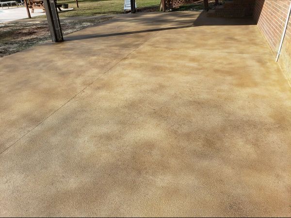 Brown, mottled concrete patio with shadows, possibly stained, next to a brick wall and a covered area.
