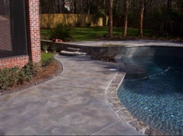 Concrete pathway curves around a pool with a stone border, brick wall, and lawn in the background.