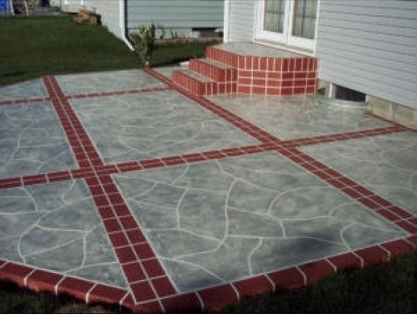 Concrete patio with red brick border and cross-shaped accents, adjacent to a house with steps.