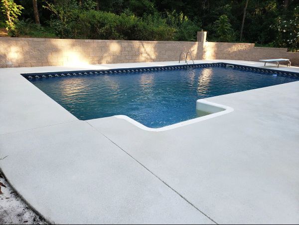 Rectangular swimming pool with light concrete deck. Blue water and trees in the background.