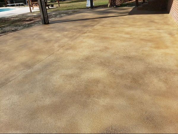Tan concrete patio with mottled color, shaded by a structure, visible near a pool and lawn.