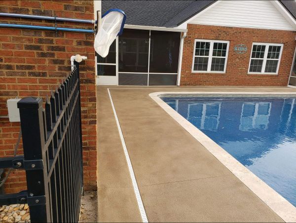 Concrete pool deck, brown with white stripe, adjacent to brick house and black metal fence.