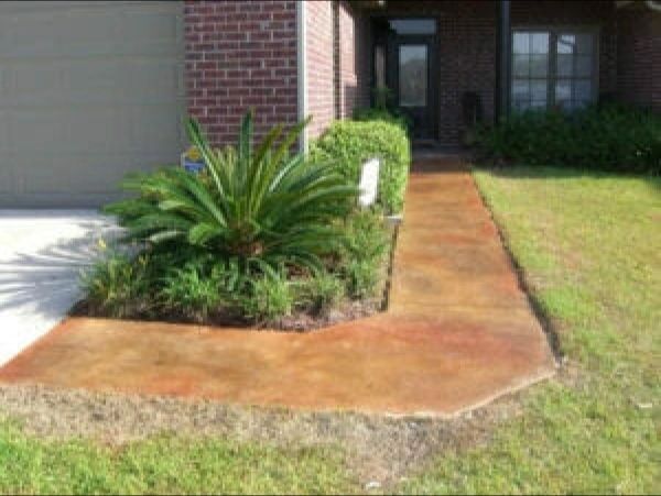 Brown stained concrete walkway leading to a house with brick and bushes.