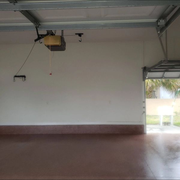Empty garage interior with a brown floor and white walls. The open garage door reveals a backyard.